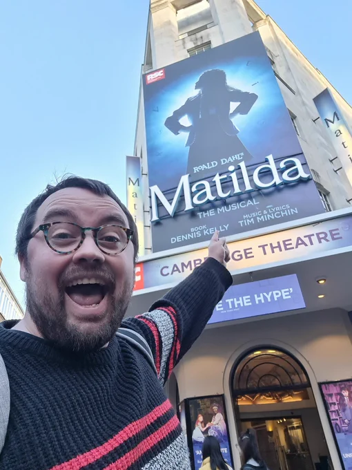 A white man, pointing excitedly at the Matilda poster outside the Cambridge Theatre