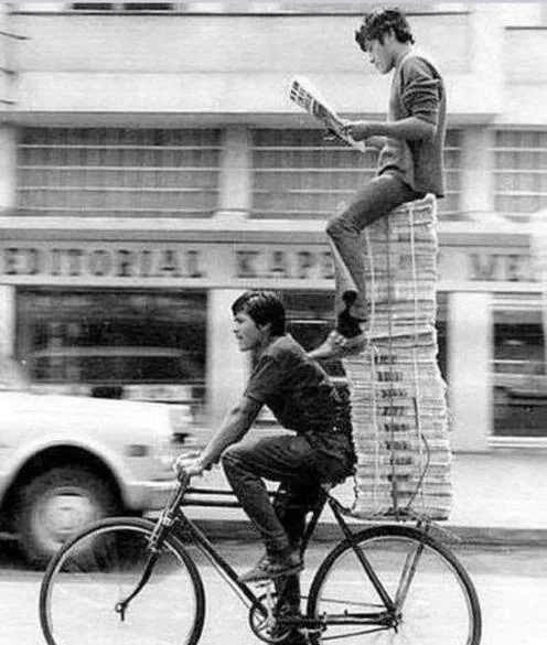 A man cycles along with a 6 foot high stack of newspapers on his back rack. Another man sits on the newspapers reading one.
Vintage image, source unknown.