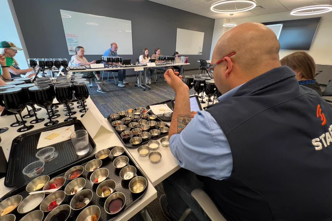 Nine individuals sit in a circle at tables covered in trays that have small containers of samples for "orienting their palates" on them. There are black goblets placed on the tables as well. Two whiteboards frame the background. 