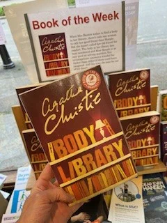  Image is a book held by a hand. The cover depicts a Bookshelf with the title of the book (The Body in the Library) spelled out in characters which look like books on the shelf.