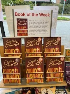 Image is a photograph of Horsley Library's Book of the Week display, a plastic stand featuring 6 copies of Agatha Christie's "The Body in the Library". Above the display is a laminated advertisement in landscape format. Said advertisement is titled: 'Book of the Week' and has a picture of the Book's cover on the left and the blurb which reads: 'When Mrs Bantry wakes to find a body in her library, there's only one woman to call: her good friend Jane Marple.  But she hasn't called her old friend for comfort.  The body in her library has been murdered and Miss Marple is so very good at solving murders.  Her investigations uncover a scandal far darker than either of them could have imagined, and this time she may be out of her depth.'