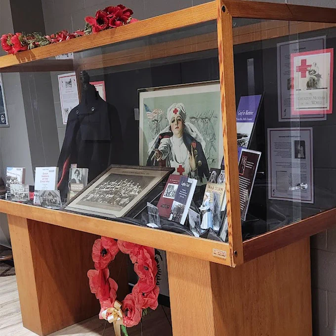 A display case featuring various historical artifacts. Inside, there are black-and-white photographs, a framed portrait of a woman in military attire, and informational brochures related to a historical event or organization. The case is decorated with poppy flowers and surrounded by additional memorabilia on the surrounding walls.