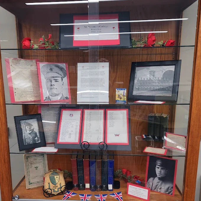 A display case featuring military memorabilia. It includes photographs of a soldier, framed documents, and historical papers, surrounded by red roses. The bottom section contains books and other artifacts, while a small British flag is partially visible at the base.