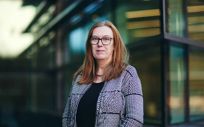Professor Sarah Gilbert wearing glasses and a patterned jacket standing in front of a modern building with large windows.