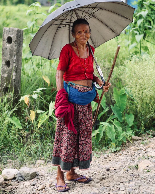 An elderly person stands on a dirt path holding an umbrella and a walking stick, dressed in traditional Nepali attire with a red top and patterned skirt.