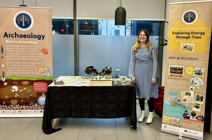 Photo of Archaeology Scotland’s Learning Assistant standing next to a display table. With 2 pop up banners on either side.