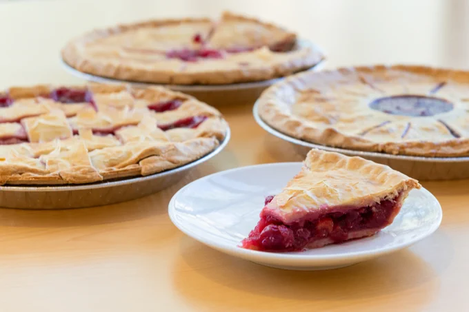 A slice of cherry pie on a white plate. Three whole pies sit on the table in the background.