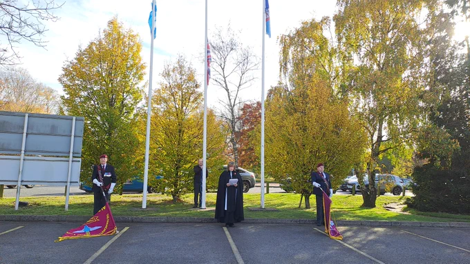 Four individuals in formal attire participate in a flag-raising ceremony outside on a sunny day, with trees and flags in the background.