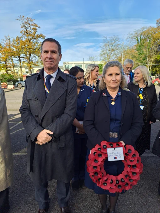 Two individuals stand side by side at an outdoor event, each wearing a poppy wreath for remembrance. The person on the left is in a dark coat, and the person on the right is wearing a badge with a lanyard. There are other attendees in the background.