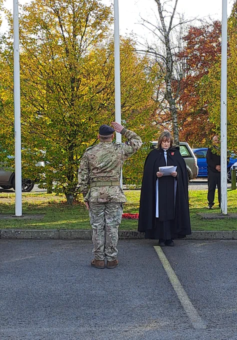 A person in military uniform saluting during the service for Armistice Day towards the flagpole and poppy wreath. Another person in a black robe holding a paper stand by flagpoles during an outdoor ceremony.