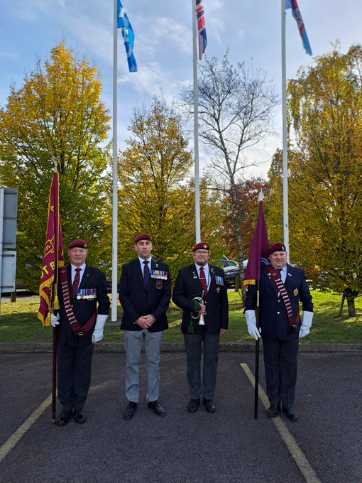 Four individuals in ceremonial attire stand solemnly outside next to trees with autumn foliage. 