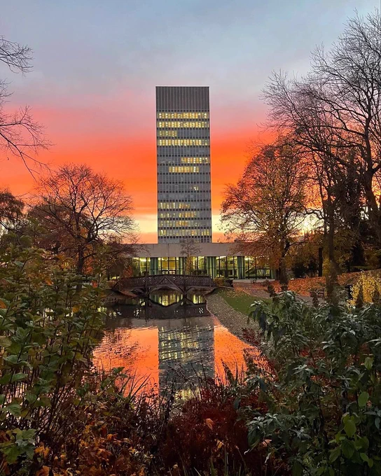 Sheffield’s iconic landmark the Arts Tower shown against a band of red sunrise sky. The building is viewed from leafy Weston Park with autumn foliage and a reflection of the building in the ornamental lake