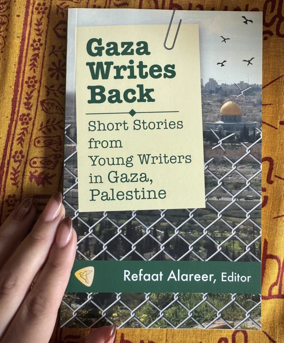 A photo of the book, Gaza Writes Back edited by Refaat Alareer on a yellow cloth background with my hand to the left.
