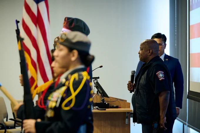 RFU campus safety officer speaks behind the podium. Students of the Waukegan High School JROTC Color Guard stand ahead but the camera has blurred their faces.
