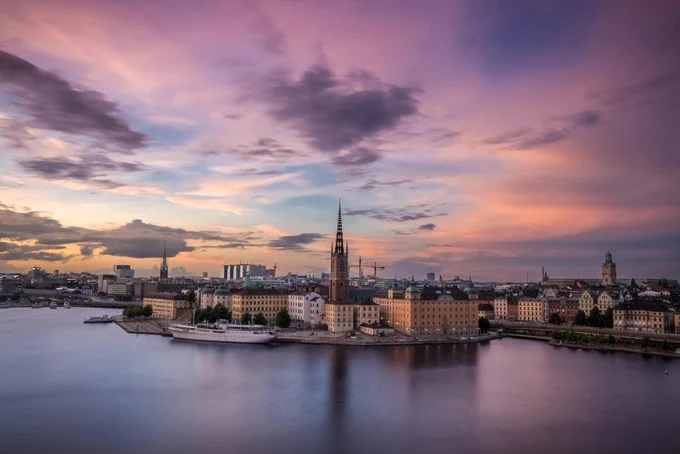 Image of Stockholm, Sweden taken from the water looking towards buildings. Photo was taken around dusk or dawn with a purple/pink/blue sky with scattered clouds. 