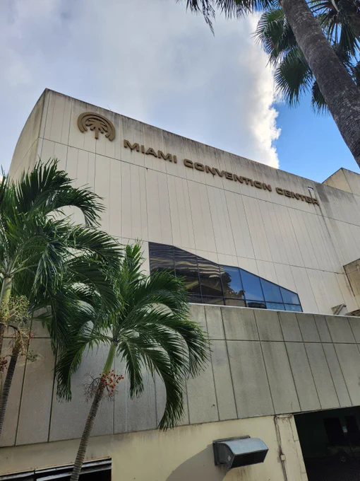 An external picture of the Miami Convention Center, with palm trees visible at the bottom left and top right corners.
