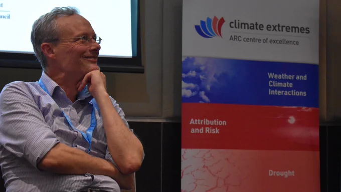 Andy Pitman is pictured wearing a striped pale shirt and glasses, sitting on a chair on a stage in front of a banner displaying the Climate Extremes logo.