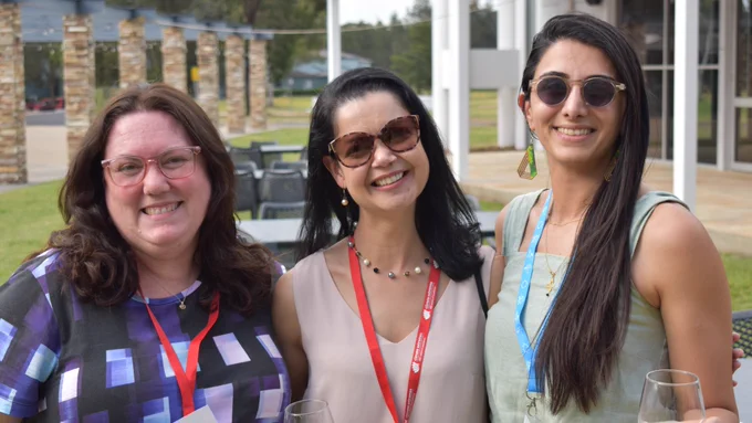 Three women are pictured outside, facing the camera and smiling. Two are wearing sunglasses. 