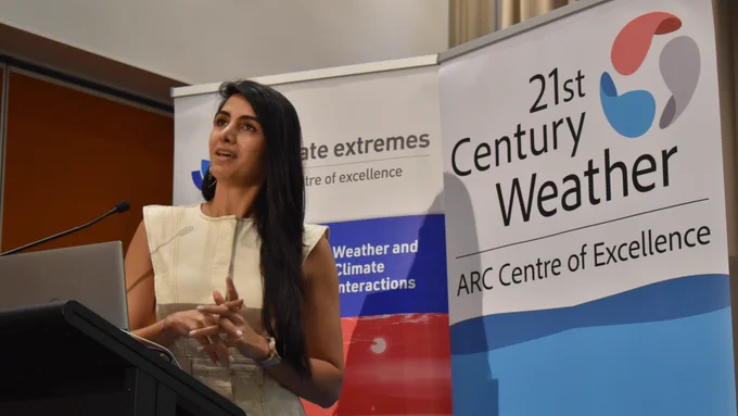 Negin Nazarian, a female researcher with dark hair, is wearing a cream dress and speaking at a lectern in front of two banners displaying the logo of Climate Extremes and 21st Century Weather.