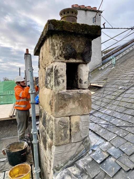 The rugged rock faced blonde sandstone on this Edwardian tenement chimney is in an aged and weathered condition. The chimney also badly needs repointed with large gaps between the stones. With one stone removed the flues inside are revealed. There are loose slates on the roof to the side. A construction worker wearing orange safety gear is slaistering a new line coating over the stonework which will help buy the chimney some time before it needs completely rebuilt in stone.