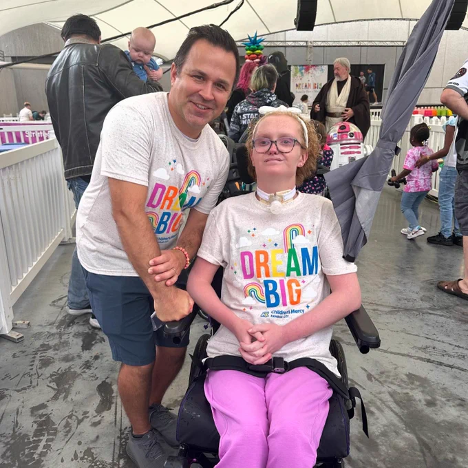 Two individuals at a 'Dream Big' event, wearing matching t-shirts with the slogan 'Dream Big'. One person is standing while holding hands with the other, who is seated in a wheelchair. They are both smiling, surrounded by a crowd and festive decorations in the background.
