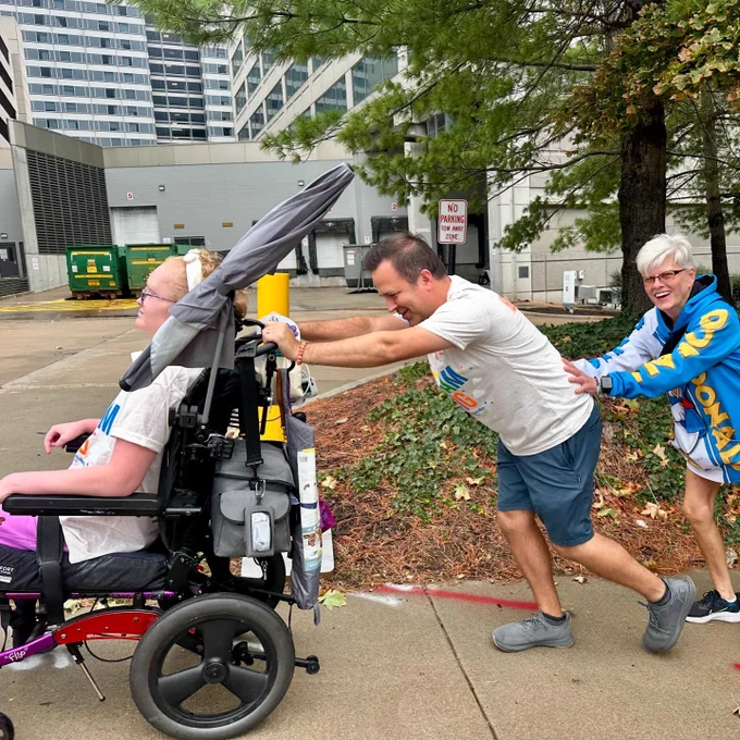 Two individuals assisting another person in a wheelchair outdoors, with a building in the background.