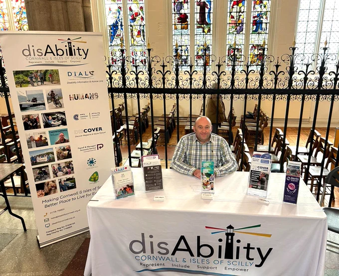 A picture of our information and advice adviser sitting by a stall with a tablecloth and leaflets. The backdrop shows a cathedral and cathedral glass. 