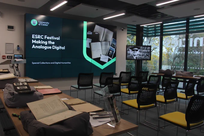 Digital Humanities seminar room set up with chairs facing a digital presentation on a large screen, and archive materials displayed on tables on both sides of the room. The title on the screen reads: 'ESRC Festival Making the Analogue Digital'.