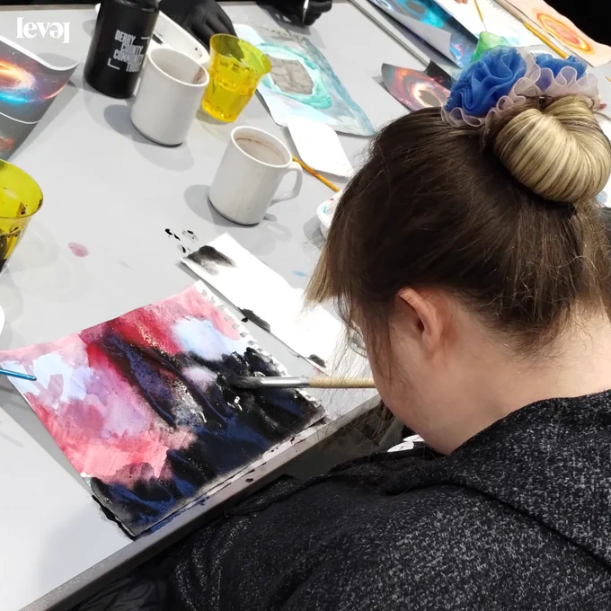 A person sits at a desk doing and abstract painting in pinks, purples, whites and blacks. The have their hair up in a bun and wear a black top.