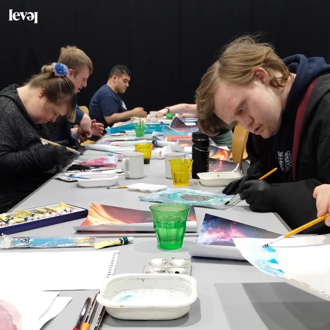A group of people sit at a long desk in a room with black walls. They are all painting and drawing. There are lots of art materials scattered on the desk.