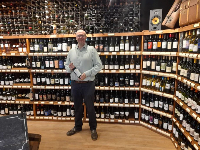 man with glasses standing in a wine shop holding a bottle of red wine 