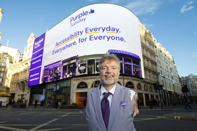 A photo of Mike standing in front of Piccadilly Lights in London. He is wearing a purple suit and tie. On the big screen is the Purple Tuesday production showing the Purple Tuesday logo and the key message 'Accessibility Everyday. Everywhere. For Everyone'. 