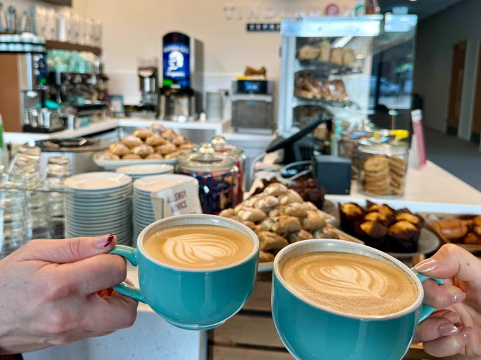 Two blue coffee cups with latte art are held in front of the counter, with sweet treats out of focus in the background at the new Tinderbox in the Adam Smith Building 