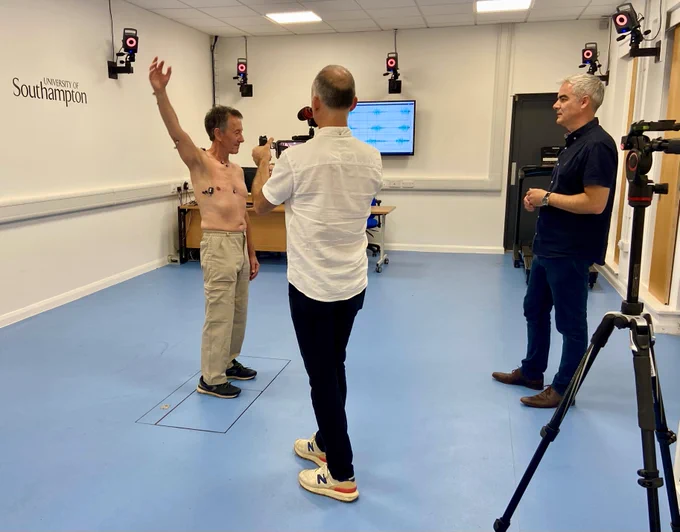 A man in white shirt and black trousers filming a man with sensors on his upper body reaching up with his right arm. A man in a blue shirt and jeans is watching, and a camera tripod is in the foreground.