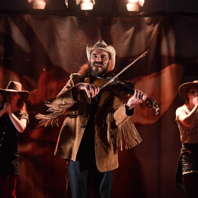 A man dressed in a cowboy hat and fringed jacket plays the violin under stage lights. His expression is joyful, and two other performers in western attire stand in the background. 
