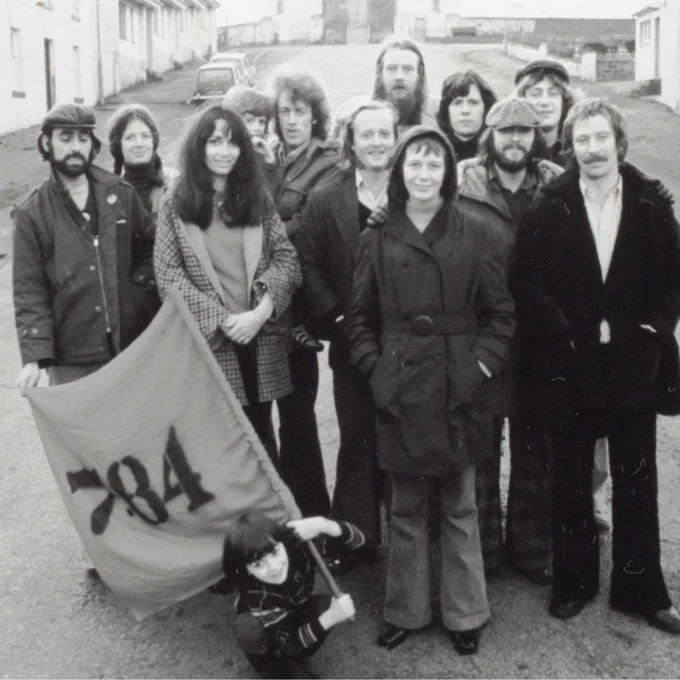 A black and white photo of a group of actors stand in the middle of the street in front of the Round Church in Bowmore. At the front of the group, a child is holding a flag that reads ‘7:84’.