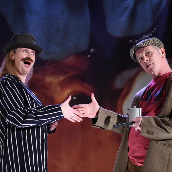 Two performers on stage shake hands in an exaggerated manner. The person on the left wears a striped suit, bowler hat, and fake moustache, while the other holds a flask and wears a tweed jacket and flat cap. 