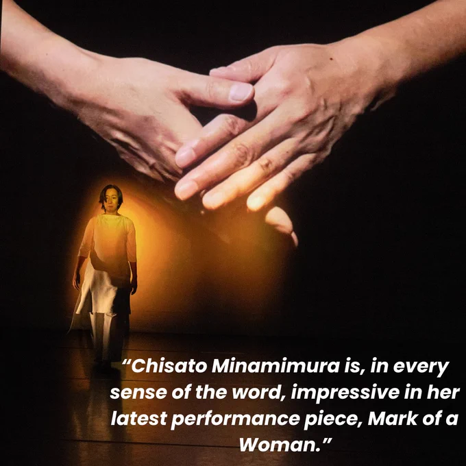 A performance image of Chisato Minamimura performing Mark of A Woman. Chisato wears a beige sleeveless dress with a beige long sleeve t-shirt beneath. Chisato is performing, walking on the stage. There is a projection behind her of lots of a set of hands holding one one another. There is text on the image, which says: “Chisato Minamimura is, in every sense of the word, impressive in her latest performance piece, Mark of a Woman.”