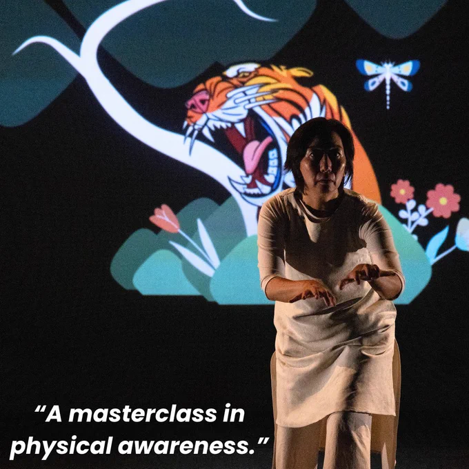 A performance image of Chisato Minamimura performing Mark of A Woman. Chisato wears a beige sleeveless dress with a beige long sleeve t-shirt beneath. Chisato is performing, crouched over, there is a tiger projection behind her with flowers around it. There is text on the image, which says: “A masterclass in physical awareness.”