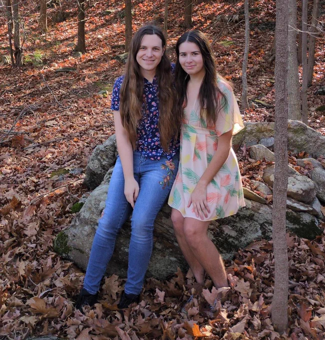 Photo of Celeste and Cara sitting together on a rock in the forest. The season is fall and leaves from the surrounding trees have fallen to the ground. Both are smiling and have long dark hair. Celeste is wearing a shirt and jeans with floral patterns. Cara is wearing a dress with floral patterns.