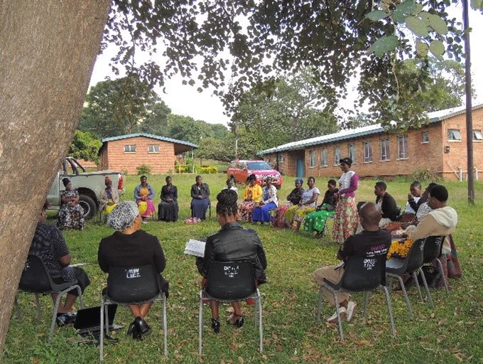 A group of women sitting on chairs. They are forming a circle. Inside the circle is a woman standing and she is speaking to the group.