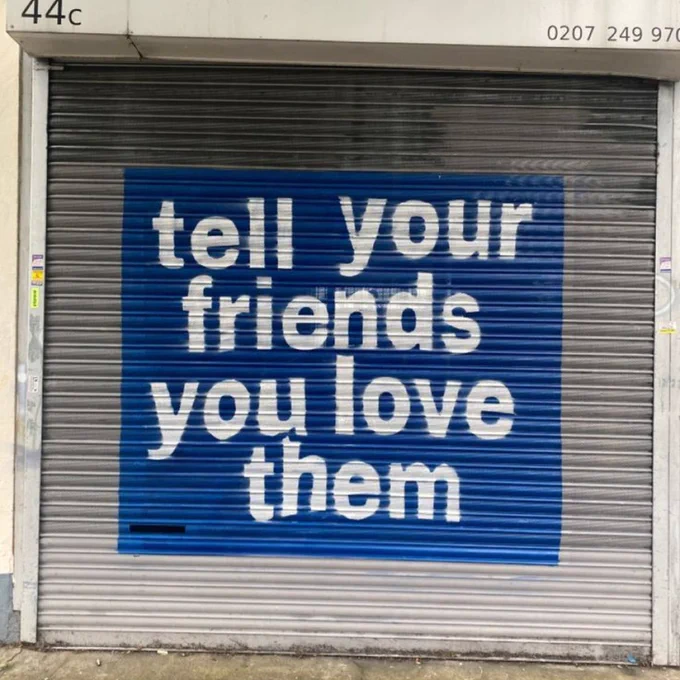 White text on a blue background saying 'tell your friends you love them' painted on a metal shutter.