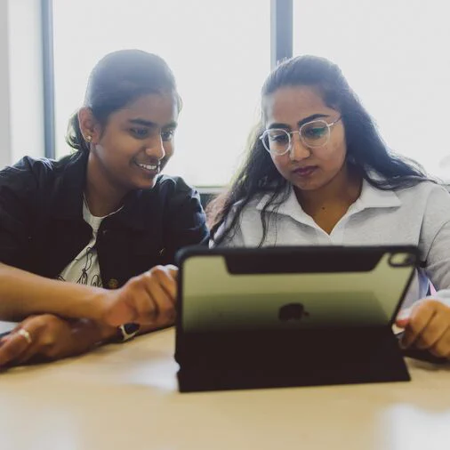 Two students are sitting side-by-side at a table looking at the screen of a propped-up tablet.