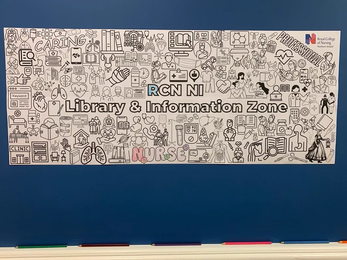 Photo of blue wall with large colouring in poster (featuring small black and white graphics of nursing, anatomy, health, and library icons with centred text: 'RCN NI Library & Information Zone), white ledge underneath with coloured pencils sitting on sill.