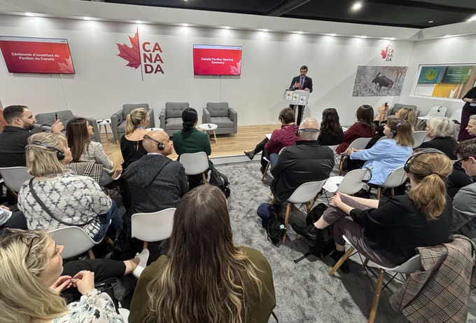 Opening ceremony of the Canadian pavilion, with an audience seated in rows facing a speaker at a podium. Behind the speaker is a wall with the Canada logo and screens displaying the event title in both English and French. 