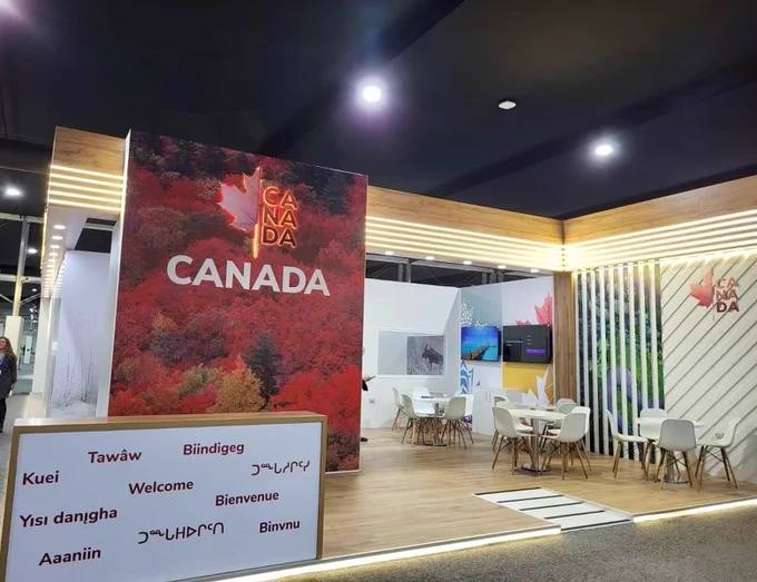 The Canadian pavilion at COP29, with a welcoming structure featuring the word "Canada" and a large image of a forest in autumn colors. A panel in the foreground displays "Welcome" in multiple languages, including several Indigenous languages.