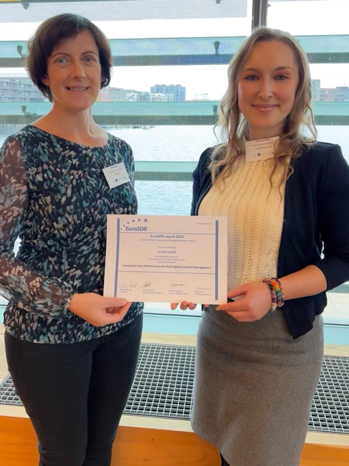 Two individuals posing with a certificate inside a building with a large window and a view of water outside.