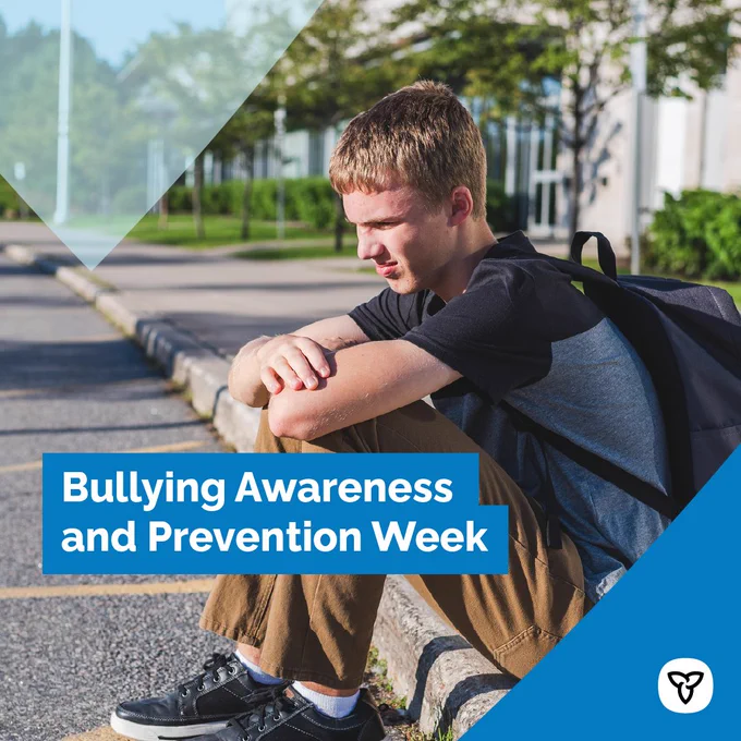A teenage boy sits by himself on a curb outside a school. Text reads: Bullying Awareness and Prevention Week