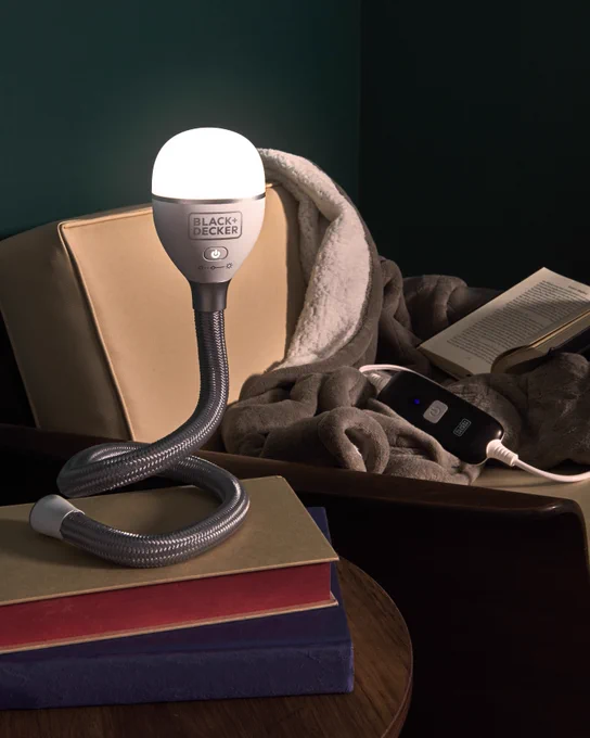 BLACK DECKER SnakeLight® light  on top of a stack of books on a table, in front of a BLACK AND DECKER heated blanket in a cozy scene.