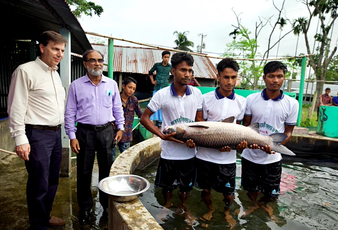 Dr. Mark Lawrence and Dr. Md. Rafiqul Islam Sarder observe three men holding a large broodstock carp in an aquaculture tank at a fish hatchery in Bangladesh